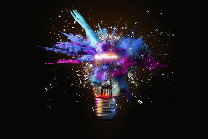 Colorful explosion of blue, purple, and pink powder bursting from a glowing light bulb against a black background.