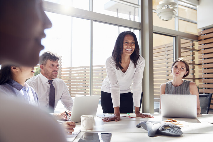 A diverse group in a meeting, with a woman standing, smiling at her colleagues.