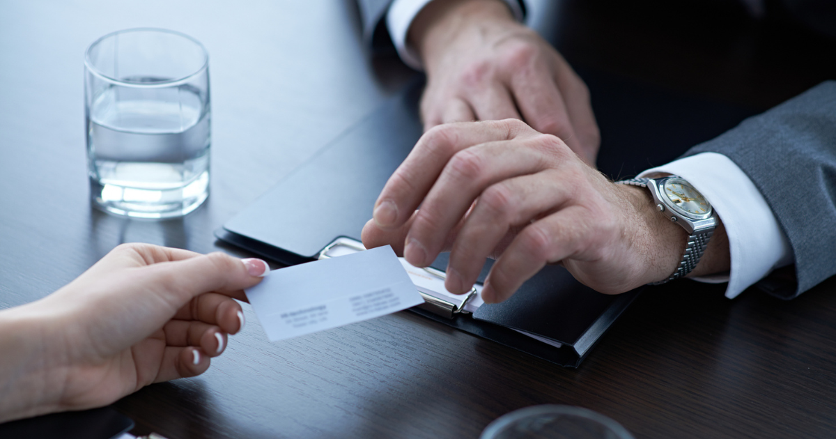 Person handing business card to professional across desk with laptop and glass of water