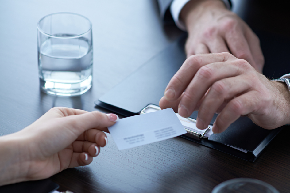 Person handing business card to professional across desk with laptop and glass of water