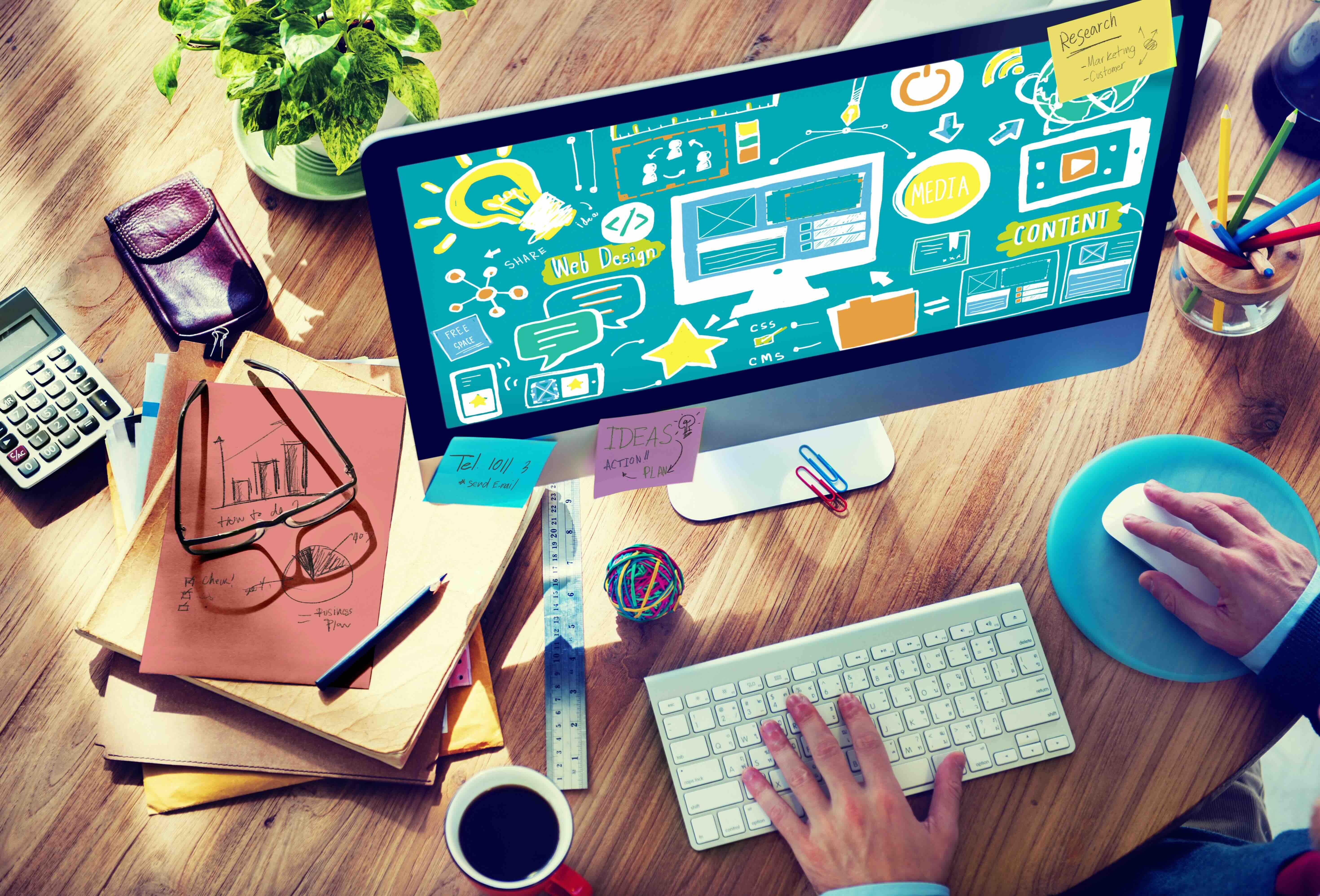 Designer workspace with computer displaying web design graphics, keyboard, notes, and office supplies on wooden desk