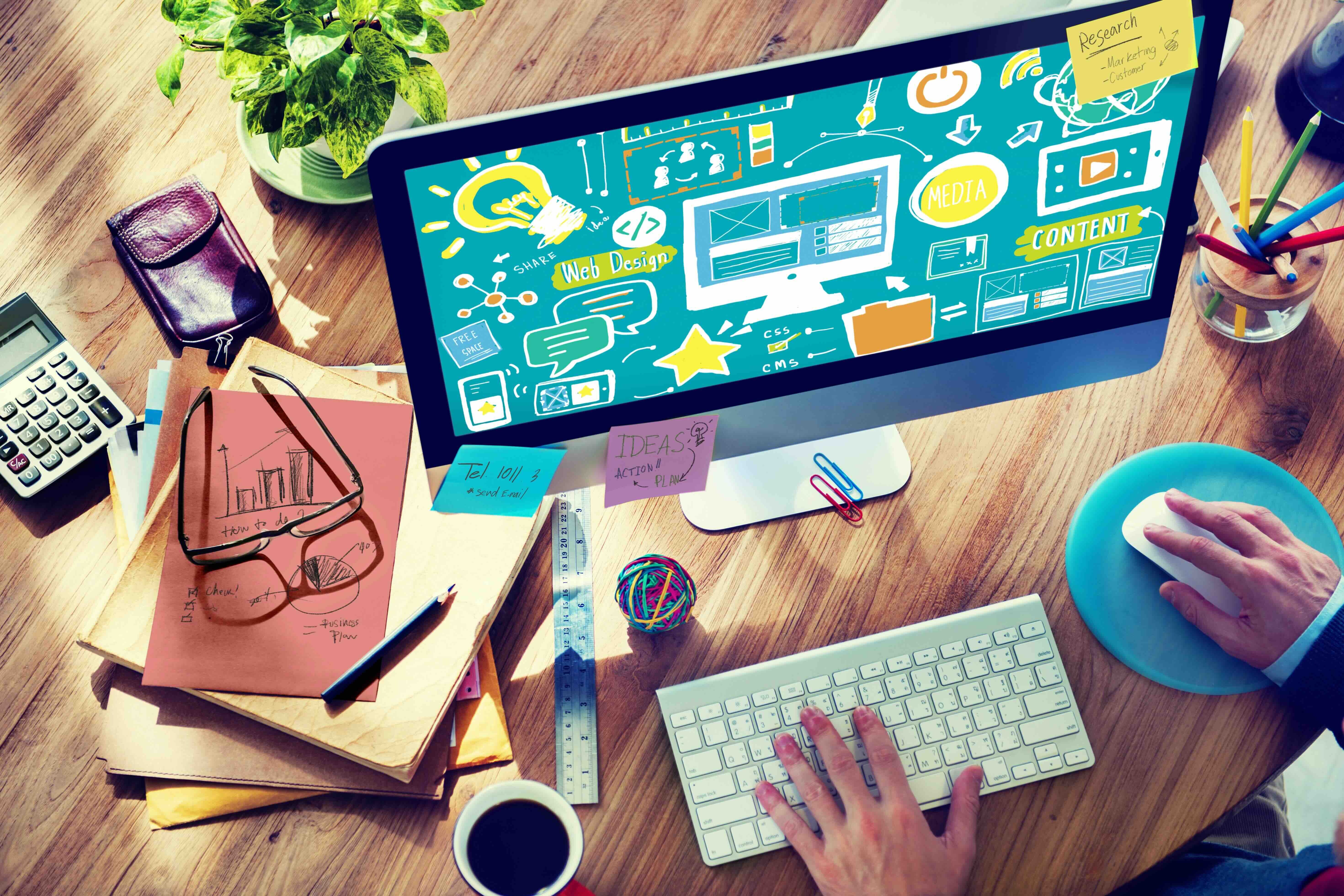 Designer workspace with computer displaying web design graphics, keyboard, notes, and office supplies on wooden desk