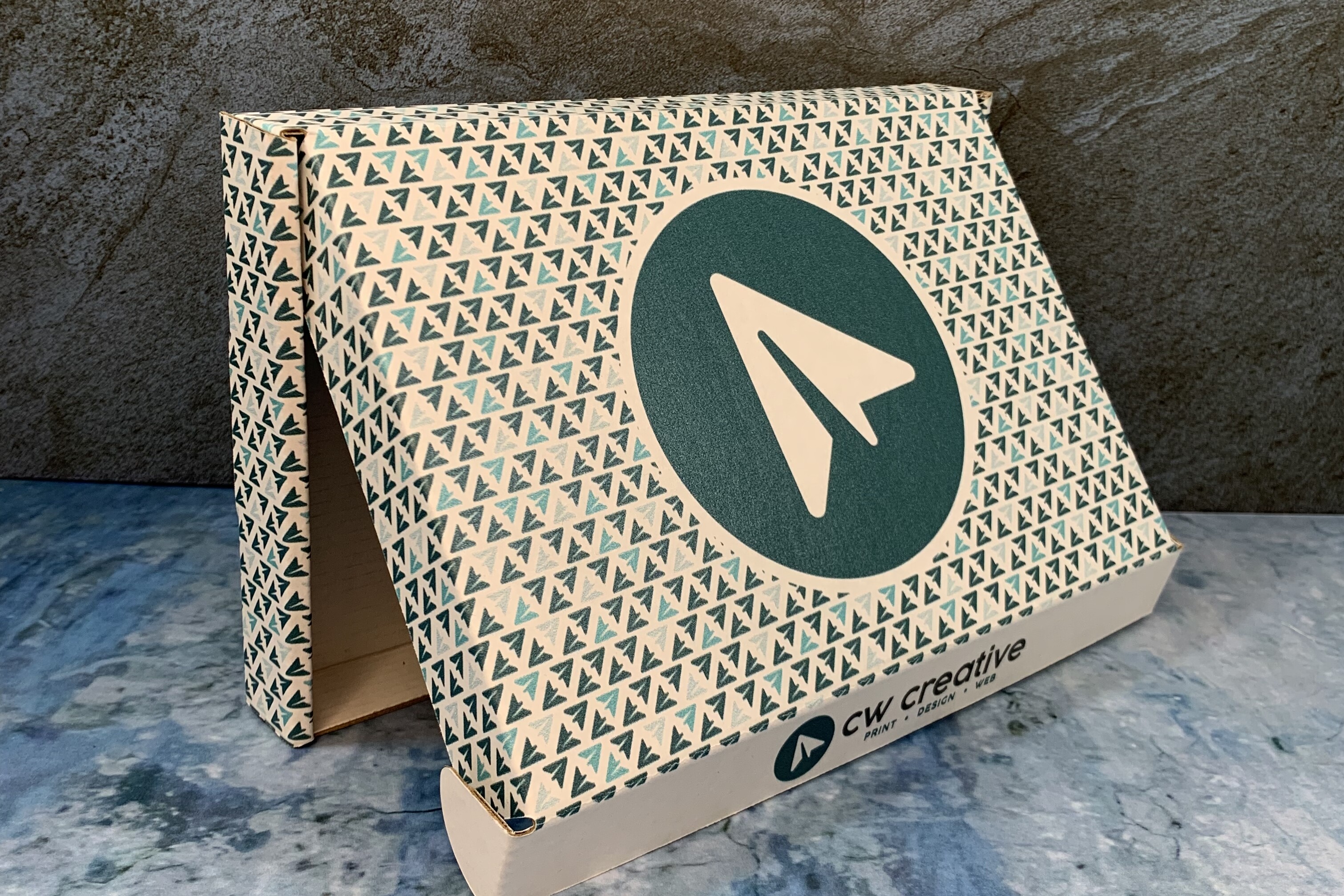 Folded display box with teal arrow pattern and CW Creative logo on a marble surface.