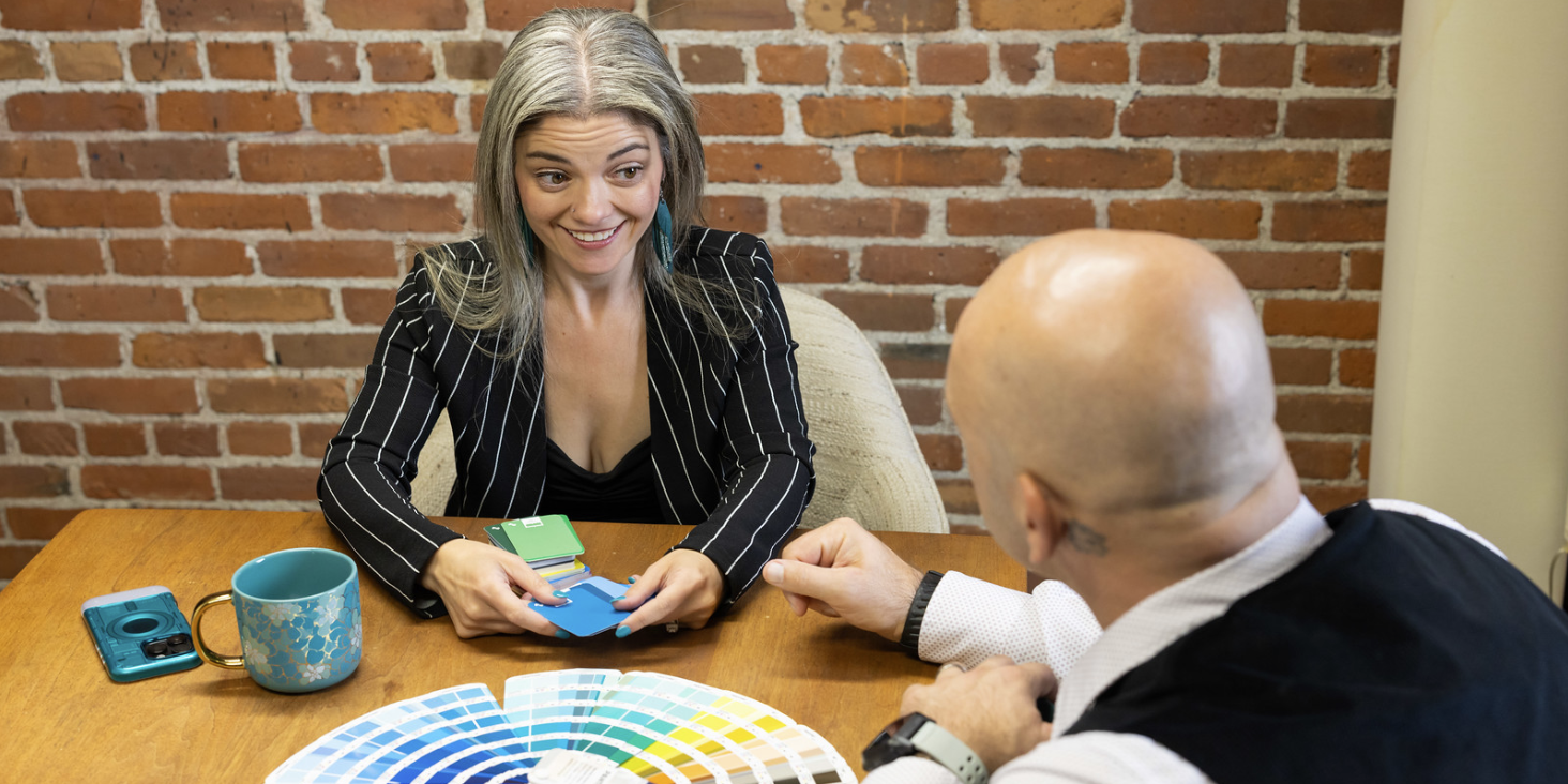 Two people discuss color samples at a table with a color swatch book and a coffee mug.