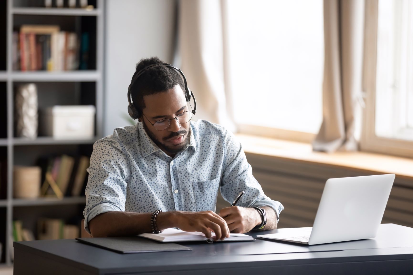young man at a laptop wearing headphones and writing on a notepad