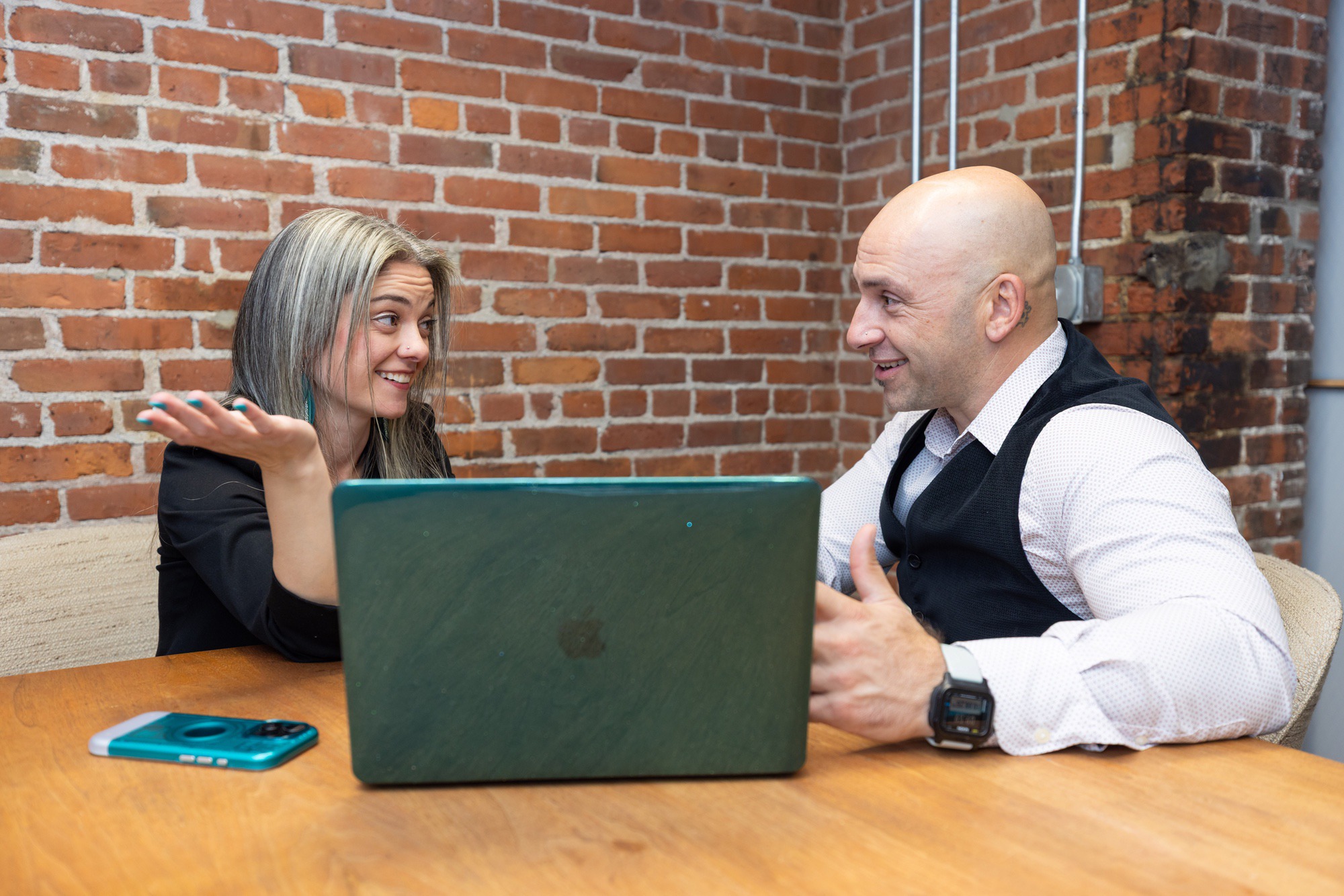 Two people engaged in a conversation over a laptop in a brick-wall setting.